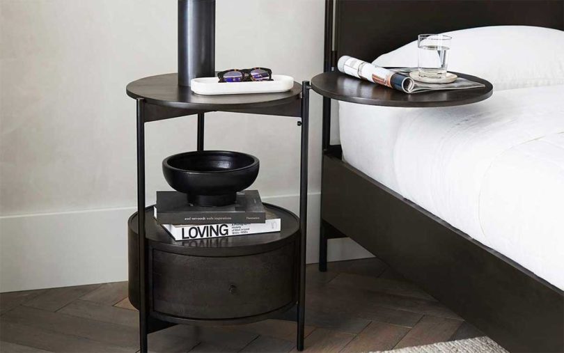 una mesa auxiliar negra con un estante redondo en la parte inferior al lado de la cama